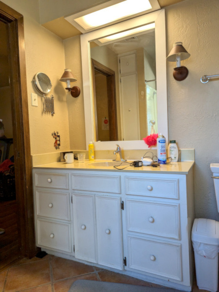 Image of bathroom before remodel. Image shows a white vanity, beige countertop, beige walls, fluorescent light , red floor tile