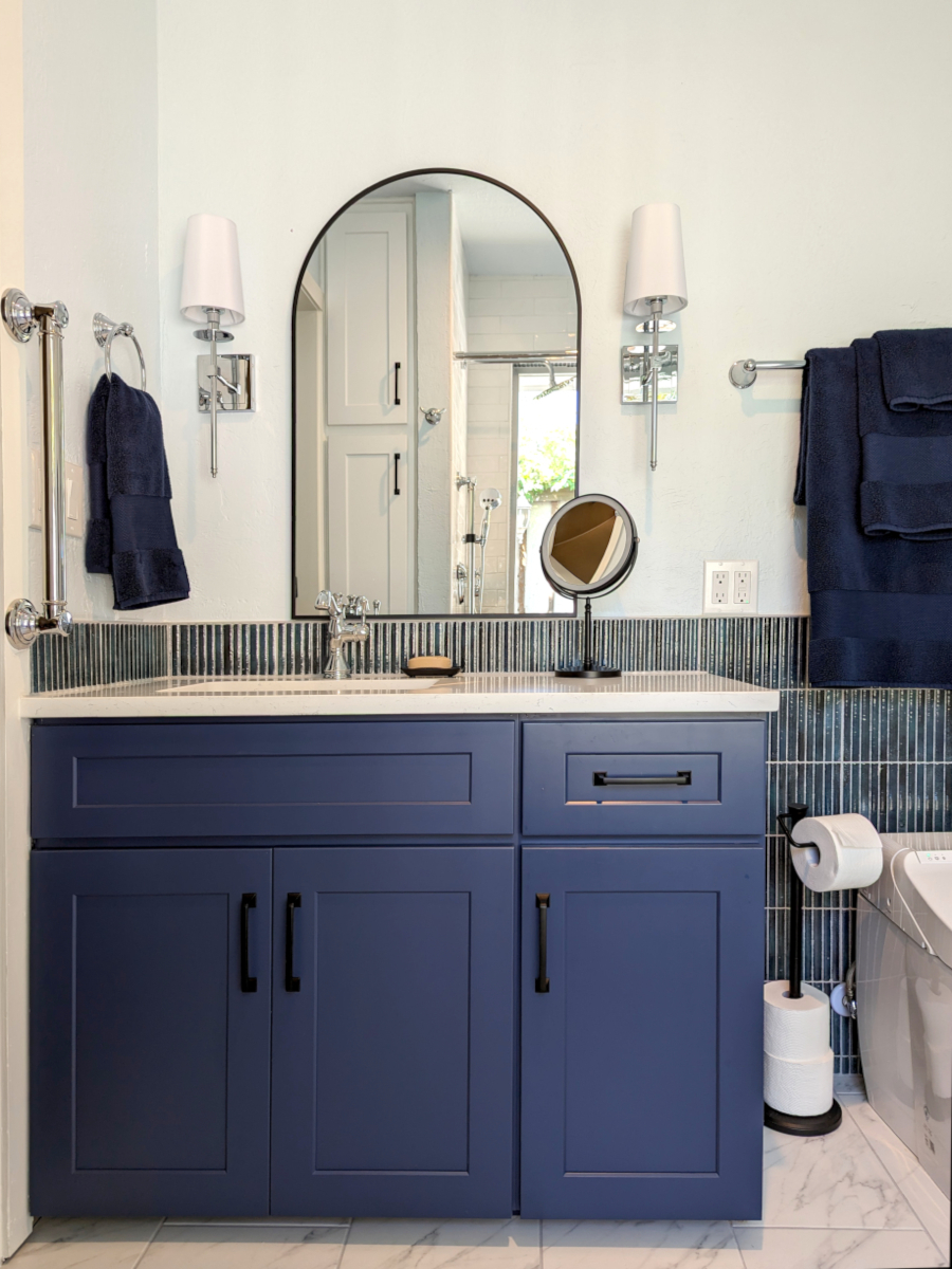 bathroom remodel after image. The image shows a shaker-style blue vanity with a quartz countertop, elegant chrome fixtures, black mirror and blue accent backsplash